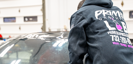 Person wearing a PRIME Graphics Lab hoodie working on the windshield or roof of a car inside a garage. The hoodie displays the company logo, contact number, and services like wraps and tinting in bold white and pink text.