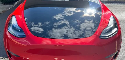 Close-up of a shiny red Tesla with a highly reflective hood that mirrors a blue sky filled with clouds and sunlight, showcasing a pristine, glossy finish.