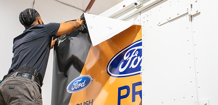 A person installs a large vinyl wrap on the back of a white truck, partially covering it with a design that features the Ford logo and the word “PRO” in bold letters.