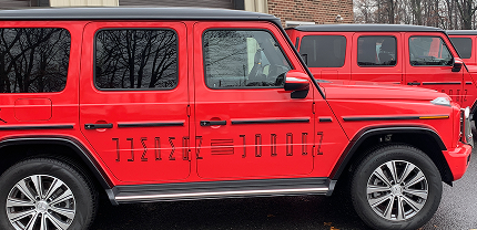 A close-up of a bright red Mercedes-Benz G-Class SUV parked beside two identical models. The SUV has unique horizontal black accents on the doors and chrome detailing on the wheels, with reflections of bare trees visible in the windows.