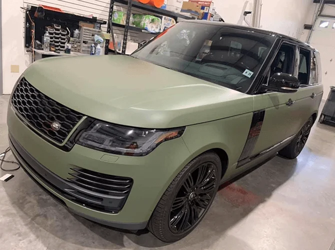 A matte army green Range Rover SUV parked inside a garage or workshop. The vehicle has black trim and wheels, with detailing that gives it a sleek, custom appearance. A workbench and shelves with tools and supplies are visible in the background.