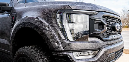 Close-up of the front left side of a Ford Black Widow truck wrapped in a pixelated digital camo vinyl. The image highlights the rugged fender, custom headlight design, and aggressive all-terrain tires.