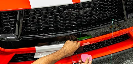 Close-up of a red Ford Mustang with white racing stripes as a person installs or removes a vinyl wrap using string and a pink squeegee tool. The iconic Mustang emblem is centered on the black honeycomb grille.