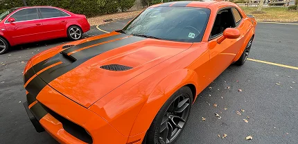 Bright orange Dodge Challenger with matte black racing stripes parked in a lot. The car features a widebody kit, aggressive hood vents, and black rims, with a red sedan visible in the background.