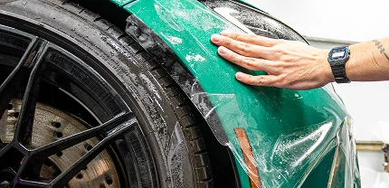 Close-up of a person's hand applying a clear protective film to the front fender of a green sports car. The image shows the glossy black wheel, tire tread, and brake disc alongside the partially adhered paint protection film.