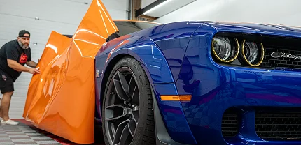 A person is applying a large sheet of glossy orange vinyl wrap to a blue Dodge Challenger in a workshop. The car features widebody fenders, custom wheels, and distinctive round headlights.