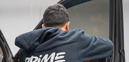 A person wearing a black hoodie with "PRIME" printed on the back is applying window tint or film to a car door window. The image is taken from behind as they work carefully inside a workshop.
