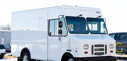 White step van with a high roof and dual front windows parked in a lot, likely used for commercial delivery or service purposes. A green and gray business sign is partially visible in the background.