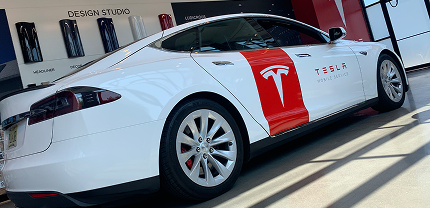 White Tesla Model S with a large red Tesla logo decal on the side parked inside a showroom. The design studio wall in the background displays various vehicle color samples and wheel options.