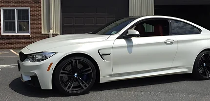 White BMW coupe with black rims parked in front of a brick building and garage. The car features a sleek, sporty design with red interior accents visible through the open window.