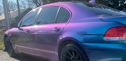 A BMW sedan with a striking color-shift vinyl wrap that transitions between purple and blue hues, parked outdoors on a sunny day. The car features tinted windows and black wheels.