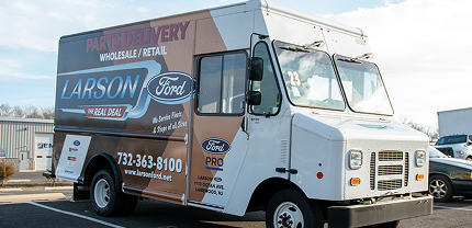 Box truck with a branded wrap for Larson Ford displaying “Parts Delivery Wholesale/Retail” and contact details. The wrap features brown, black, and white tones with the Ford logo and dealership info prominently displayed.