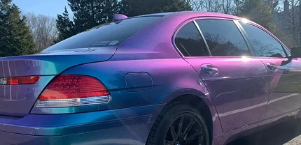 Rear-side view of a BMW sedan wrapped in a vibrant color-shifting vinyl that transitions between blue and purple. The car is parked outdoors under bright sunlight, showcasing the wrap's reflective sheen and black rims.