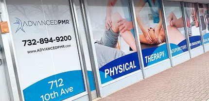 Storefront windows of Advanced PMR in New Jersey, displaying large printed graphics with people receiving physical therapy. The windows read: "PHYSICAL THERAPY," "ACUPUNCTURE," and "CHIROPRACTIC," along with the contact number 732-894-9200 and address 712 10th Ave.
