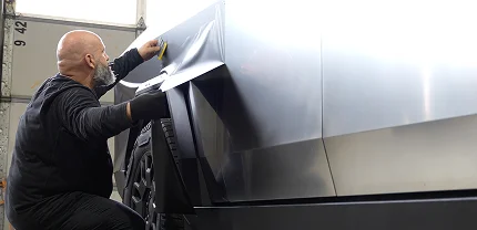 A man with a beard and black gloves applies a matte vinyl wrap to the side of a Tesla Cybertruck inside a garage, carefully smoothing out the material over the truck’s angular body panels.