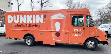 Bright orange Dunkin’ truck parked outdoors, featuring a large coffee cup graphic and slogans that read “Sipping is Believing” and “Full Steam Ahead” on the side.