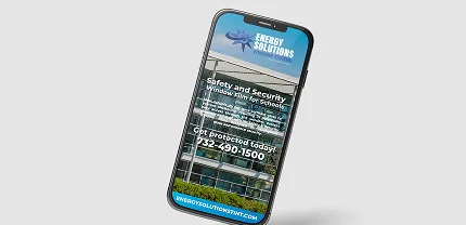 Smartphone displaying a promotional graphic for Energy Solutions Window Tint, focused on safety and security window film for schools. The ad includes a building image and reads: “Safety and Security Window film for schools,” followed by a phone number, 732-490-1500, and a website, energysolutionswt.com.