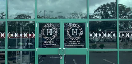 Front glass doors of Hooked Boxing & Fitness with black circular logos featuring a white "H" in the center. The left door reads “HOOKED Boxing & Fitness,” and the right door includes the phone number 732-637-7011 and “By Appointment Only.” Decorative black and red geometric patterns stretch across the surrounding windows.