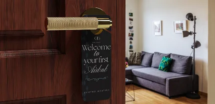 A wooden door with a gold handle and a black door hanger that reads, “Welcome to your first Airbnb – Sweetest Staycation.” In the background, a cozy living room with a gray sectional couch, green throw pillow, floor lamp, and wall art is visible.