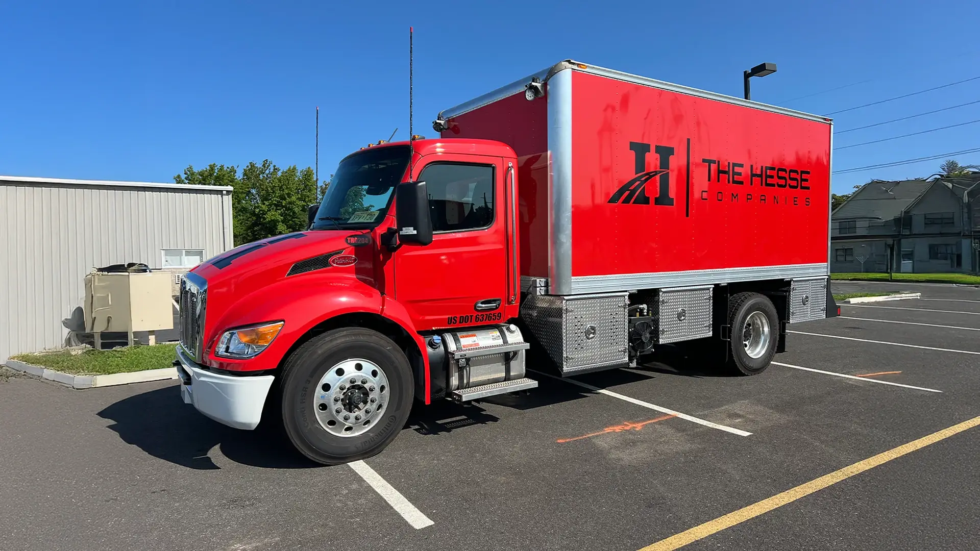 A bright red box truck branded with "The Hesse Companies" logo is parked in a lot under clear blue skies. The truck has a Peterbilt badge, chrome detailing, and is marked with a US DOT number on the cab.