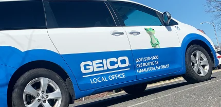 GEICO-branded van with a white and blue color scheme, parked in a lot. The van features the GEICO gecko mascot and contact information for the local office in Hamilton, NJ, including phone number and address.