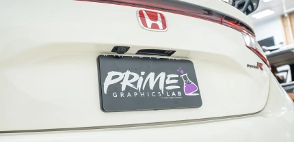 Close-up of the rear of a white Honda Type R with a custom license plate that reads "PRIME GRAPHICS LAB" in bold lettering, featuring a purple beaker logo on the right.