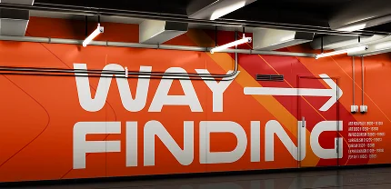 Bold orange wall mural inside a building with large white text reading “WAY FINDING” and a right-pointing arrow. The background includes diagonal red and yellow accents and a list of directional locations in small white text.
