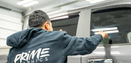 A person wearing a "PRIME GRAPHICS" hoodie is applying or inspecting tint or wrap on the rear window of a dark-colored SUV inside a brightly lit garage.