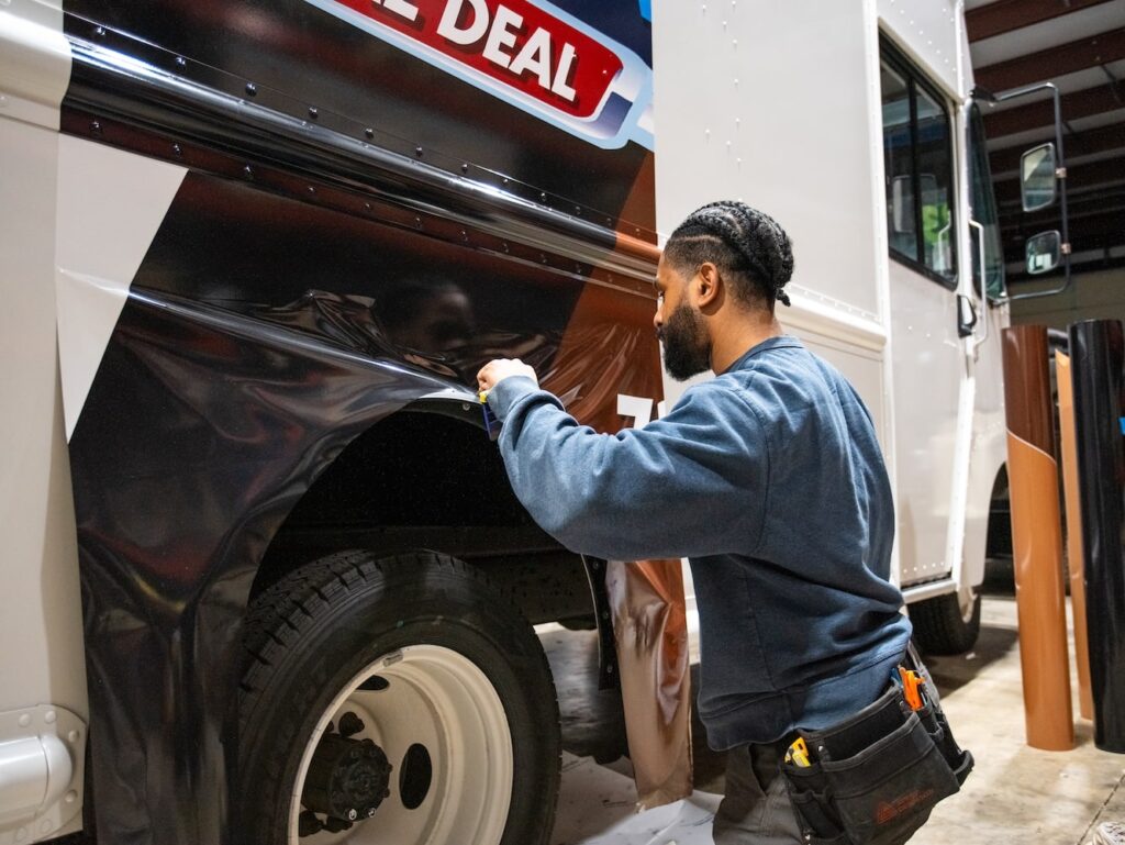 Employee at Prime Graphics Lab carefully applying vinyl wrap to the side of a large commercial truck in a workshop.